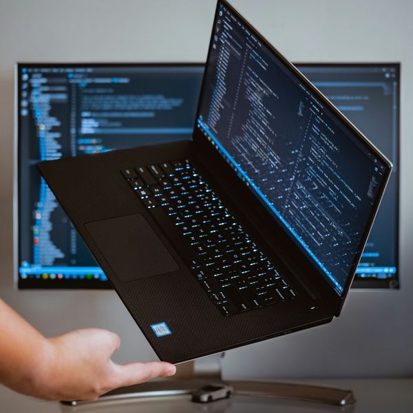 Eine Person hält einen modernen Laptop mit einem geöffneten Code-Editor vor einem Monitor, der ebenfalls Code anzeigt.